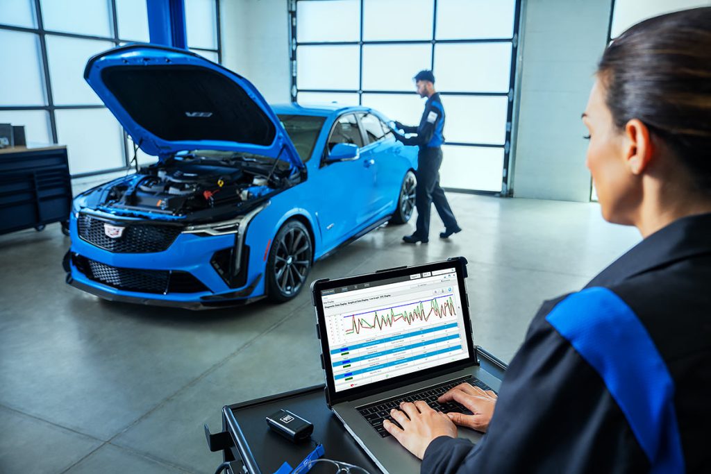 A technician uses a laptop to analyze vehicle diagnostics while another technician inspects a blue performance sedan with its hood open inside a service bay.